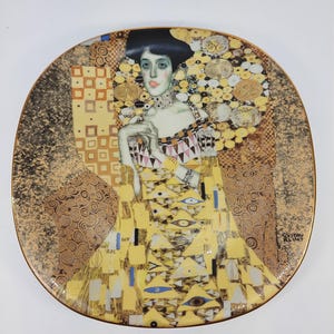 May include: A decorative plate featuring a portrait of a woman in a yellow dress with geometric patterns, reminiscent of Gustav Klimt's art. The plate has a gold rim and a detailed background with gold and brown tones.