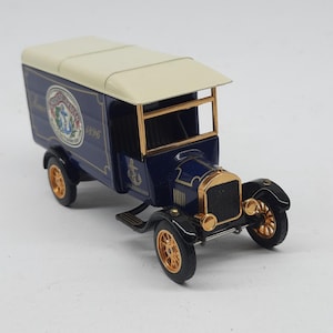 May include: A die-cast metal model delivery truck featuring a dark blue body, a cream-colored roof, and gold-toned wheels and trim. The side panel displays a circular logo with an anchor and the text "San Francisco 1896" in white and red.