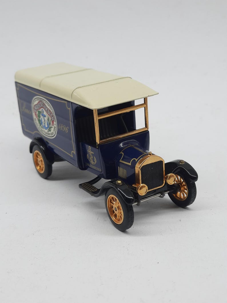 May include: A die-cast metal model delivery truck featuring a dark blue body, a cream-colored roof, and gold-toned wheels and trim. The side panel displays a circular logo with an anchor and the text "San Francisco 1896" in white and red.