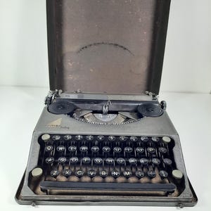 May include: A gray typewriter with a black keyboard and the word "rmebaby" printed on the front. The typewriter is open and the lid is propped up behind it.