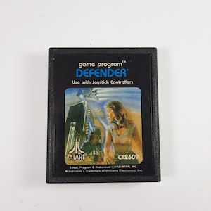 Può includere: Cartuccia di gioco Atari Defender nera. L'etichetta presenta il titolo del gioco, "DEFENDER", e una grafica colorata di una donna e una scena futuristica. La cartuccia è progettata per l'uso con i joystick.