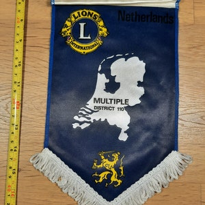 Può includere: Un pennone blu con un contorno bianco dei Paesi Bassi, il testo "Netherlands", "Lions International" e "Multiple District 110". Il pennone ha un emblema di leone giallo e una frangia bianca.