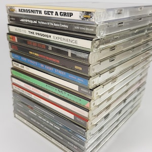Puede incluir: Una pila de CDs de música con varios títulos de álbumes, incluyendo "Aerosmith Get a Grip" y "The Prodigy Experience". Las cajas de los CDs son de plástico transparente y están apiladas.
