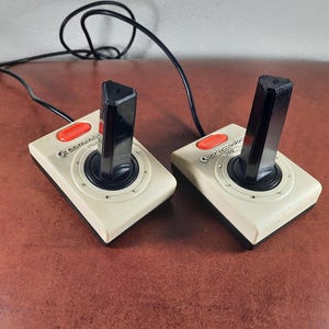 Könnte beinhalten: Zwei weiße und schwarze Atari-Joystick-Controller mit roten Knöpfen. Die Controller sind durch ein schwarzes Kabel verbunden.