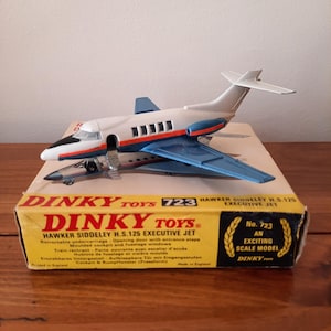 May include: A Dinky Toys Hawker Siddeley H.S.125 Executive Jet model airplane, displayed on its original box. The toy plane is white, blue, and silver, with black window details. The box is yellow and red, with text in English.