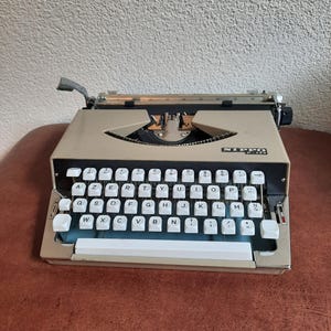 May include: A vintage beige typewriter with white keys. The keys are arranged in a QWERTY layout. The typewriter has a silver carriage return lever and the brand name "NIPPO" is visible on the front.