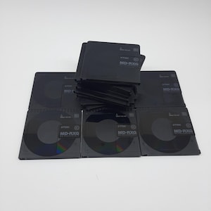 Puede incluir: Una colección de discos MiniDisc TDK MD-RXG negros y grabables. Los discos están apilados y dispuestos sobre una superficie blanca. Cada disco tiene el logotipo de TDK y MD-RXG.