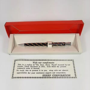Puede incluir: Una tarjeta blanca con texto negro en un borde decorativo. El texto dice "Con nuestros cumplidos. Esta es una réplica del "Lápiz Ever Sharp" inventado por nuestro fundador Tokuji Hayakawa en 1915, de donde se deriva el nombre actual de la empresa. Nos gustaría presentártelo, junto con nuestro sincero agradecimiento por tu continuo apoyo y cooperación. SHARP CORPORATION."