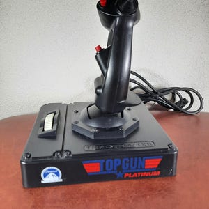 Può includere: Joystick nero Top Gun Platinum con pulsanti rossi e una base nera. La base ha un adesivo bianco con la scritta "Top Gun Platinum" in rosso, bianco e blu.