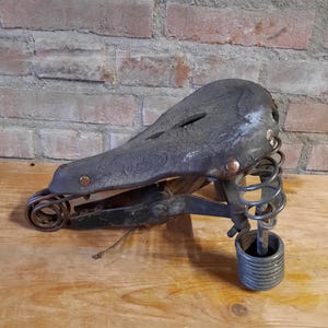 May include: A vintage black leather bicycle saddle with a spring suspension system. The saddle is worn and has a few scratches. The spring system is made of metal and has a black finish.