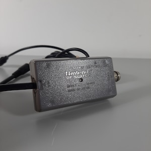 Può includere: Un interruttore RF Nintendo grigio per la console NES. L'interruttore ha un cavo nero collegato ad esso. Il testo sull'interruttore recita "Nintendo RF Switch Model No. NES-006 Made in Japan".