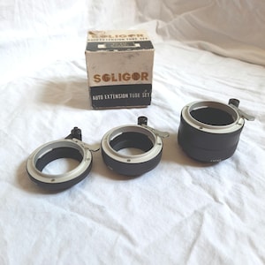 Puede incluir: Un juego de tres tubos de extensión de cámara de metal negro con detalles plateados. Los tubos están etiquetados "Soligor Auto Extension Tube Set".