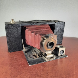 May include: A vintage black and red bellows camera with a leather exterior and a viewfinder on the side. The camera has a lens with a silver rim and a red bellows.