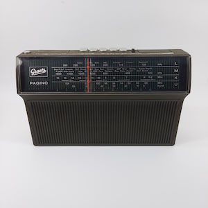 Könnte beinhalten: Ein Vintage-Radio Graetz Pagino in Schwarz und Grau. Das Radio hat einen dunkelgrauen, geriffelten Sockel und eine schwarze Oberseite mit einem Abstimmrad und Stationsanzeigen. Auf der Vorderseite des Radios sind das Graetz-Logo und das Wort "PAGINO" zu sehen.