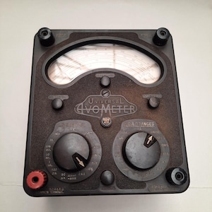 Puede incluir: Un antiguo Universal G-V-O-Meter, un dispositivo de prueba eléctrica marrón oscuro. Cuenta con dos diales analógicos, múltiples perillas y un terminal rojo. El medidor muestra varias escalas y rangos para mediciones eléctricas.