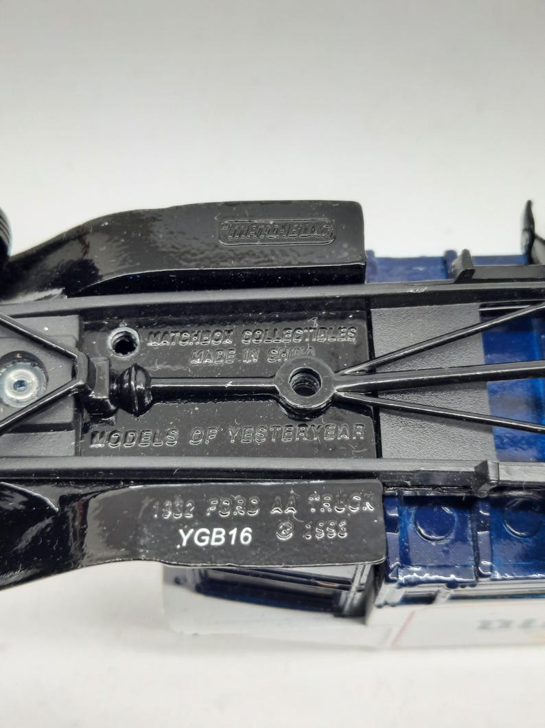 May include: The underside of a blue and black die-cast toy truck featuring the embossed text Matchbox Collectibles, Made in China, Models of Yesteryear, 1932 Ford AA Truck, and YGB16. The chassis is black metal with visible screw holes.