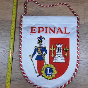 May include: White fabric pennant with red and white trim. The pennant features a red and white crest with a tower and fleur-de-lis. The text "EPINAL" is printed in red at the top of the pennant. The crest also includes a yellow and red logo with the text "LIONS INTERNATIONAL".