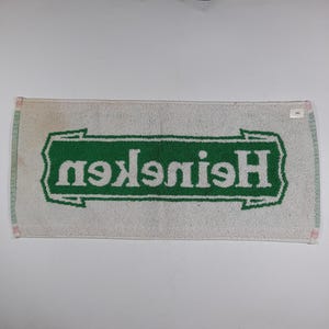 Pode incluir: Toalha retangular branca com borda verde e logotipo verde "Heineken" em fonte estilizada. A toalha apresenta sinais de desgaste.