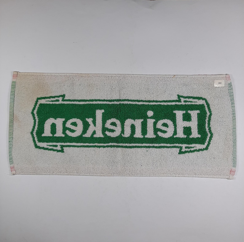 Pode incluir: Toalha retangular branca com borda verde e logotipo verde "Heineken" em fonte estilizada. A toalha apresenta sinais de desgaste.
