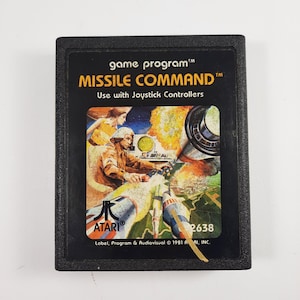 May include: Black Atari Missile Command game cartridge. The label features a colorful illustration of a military scene with the game title. The text "Use with Joystick Controllers" is also visible. The cartridge is a vintage video game.
