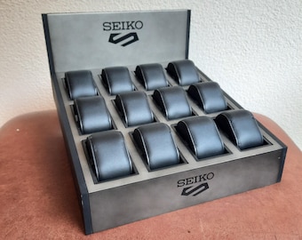 May include: A gray Seiko display stand featuring twelve black watch cushions arranged in a grid. The stand has a slanted design with the Seiko logo and emblem printed on the top back panel and the front base. The structure is made of gray material.