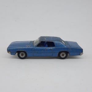 May include: A vintage blue die-cast toy car in a coupe body style. The vehicle features a metallic blue exterior finish with visible signs of wear, black wheels with silver hubs, and a clear windshield revealing a blue interior cabin structure.