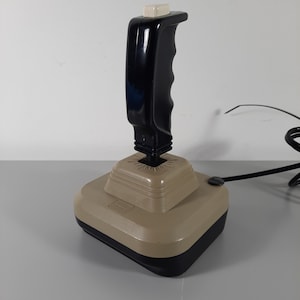 May include: A black and beige vintage joystick with a white button on the top of the handle. The joystick is connected to a cord.