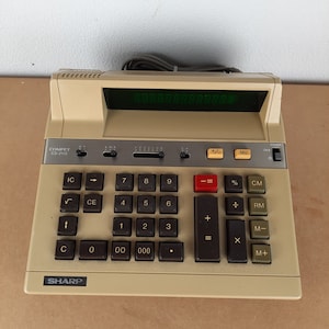 May include: A beige Sharp CS-2115 calculator with a green digital display. The calculator has a standard layout with number keys, function keys, and an equals key. The calculator is on a brown surface.