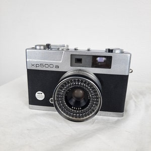 May include: A silver and black 35mm film camera with a Hexar lens. The camera has a black leatherette covering and a silver top plate. The camera has a black lens cap and a silver flash shoe.
