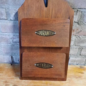 Könnte beinhalten: Ein Vintage-Wandorganizer aus Holz mit zwei Fächern. Jedes Fach hat ein Metallschild mit der Aufschrift "COURANTEN" und "WEEKBLADEN". Der Organizer ist aus dunklem Holz gefertigt und hat oben einen schwarzen Griff.