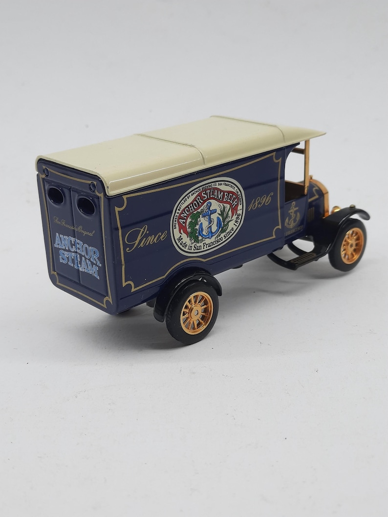May include: A die-cast model delivery truck in dark blue with a cream-colored roof. The side features the text Anchor Steam Beer, Made in San Francisco since 1896, and an anchor logo. The back displays the text Anchor Steam. It has gold-toned wheels.