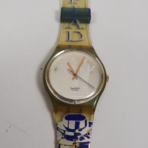 Puede incluir: Un reloj Swatch vintage con una correa amarilla transparente y una caja verde. La esfera es blanca con manecillas doradas y una ilustración detallada. La correa presenta el texto "MAD" y "12" en azul.