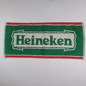 Pode incluir: Uma toalha retangular verde e branca com a palavra "Heineken" em letras brancas grandes. A toalha tem uma borda vermelha e franjas nas extremidades curtas. Provavelmente &eacute; usada para secar ou limpar.