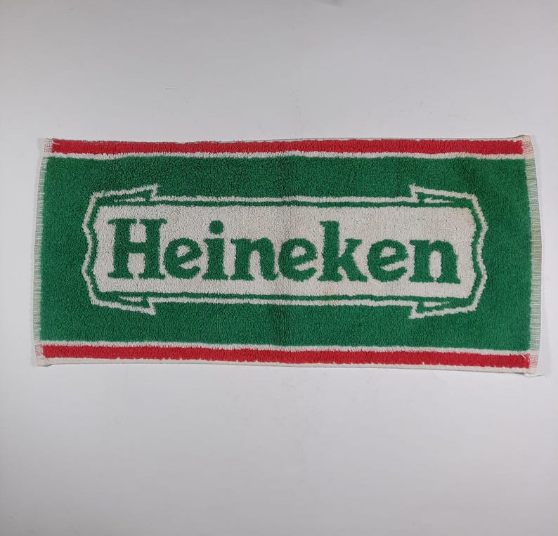 Pode incluir: Uma toalha retangular verde e branca com a palavra "Heineken" em letras brancas grandes. A toalha tem uma borda vermelha e franjas nas extremidades curtas. Provavelmente &eacute; usada para secar ou limpar.