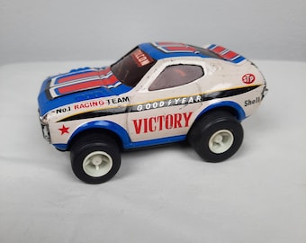 Puede incluir: Un coche de carreras de juguete vintage con un esquema de colores blanco, azul y rojo. La carrocería muestra el texto VICTORY, No.1 RACING TEAM, GOODYEAR, STP y Shell. Tiene neumáticos negros con llantas blancas y un parabrisas tintado oscuro.