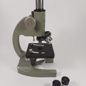 May include: An olive green vintage microscope with a black stage and two eyepieces. The microscope has a silver metal tube and adjustment knobs. The base is a rounded shape. The eyepieces are black and silver.