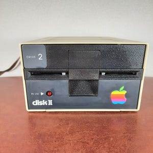 Può includere: Un'unità disco floppy Apple Disk II beige con un pannello frontale nero e un logo Apple arcobaleno. L'unità è etichettata "disk II" e "DRIVE 2".
