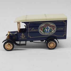 May include: A blue and cream-colored vintage delivery truck model featuring gold-toned wheels and an anchor logo. The side panel displays the text "Since 1896" and a circular emblem reading "Anchor Steam Beer, Made in San Francisco since 1896".