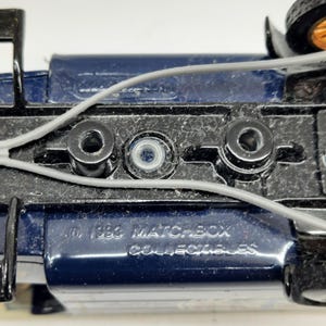 May include: The underside of a dark blue die-cast toy vehicle featuring a black chassis with three circular mounting points and thin gray plastic wiring. Embossed text on the metal surface reads 1988 MATCHBOX COLLECTIBLES in capital letters.