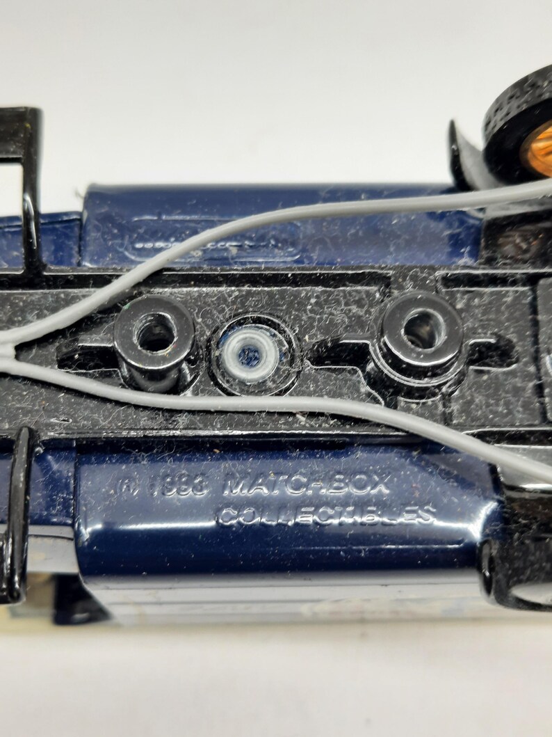 May include: The underside of a dark blue die-cast toy vehicle featuring a black chassis with three circular mounting points and thin gray plastic wiring. Embossed text on the metal surface reads 1988 MATCHBOX COLLECTIBLES in capital letters.