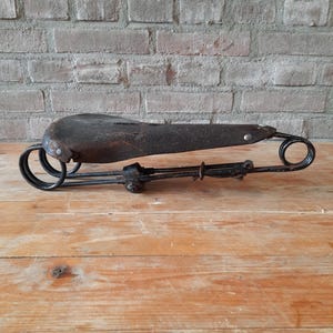 May include: A vintage bicycle saddle with a black leather seat and a metal frame. The saddle is attached to a metal spring suspension system.