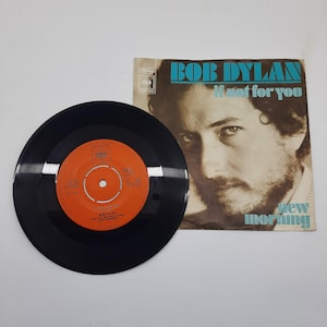 Pode incluir: Um disco de vinil preto de 45 RPM com um rótulo central laranja e uma capa de papel quadrada com um retrato em tom sépia de Bob Dylan. A capa exibe o texto BOB DYLAN, If not for you e New Morning em uma fonte azul.
