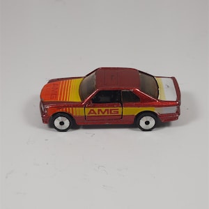 May include: A red toy car with yellow and white accents. The car has the word "AMG" printed on the side and hood. The car has black wheels and a red roof.
