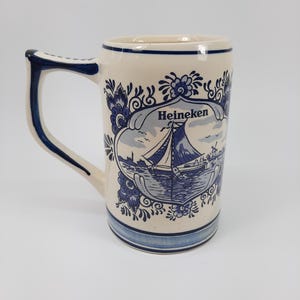 May include: Cream ceramic beer stein with blue floral and nautical design. The stein has a handle and the word "Heineken" in a banner. The design includes a sailboat on the water. The base and rim have blue stripes.