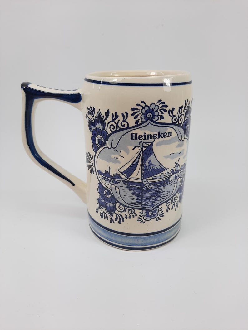May include: Cream ceramic beer stein with blue floral and nautical design. The stein has a handle and the word "Heineken" in a banner. The design includes a sailboat on the water. The base and rim have blue stripes.