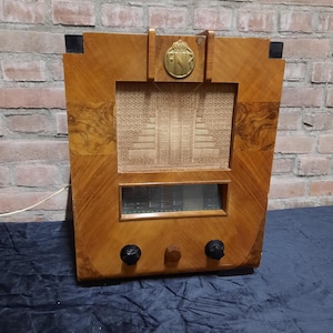 Pode incluir: Um rádio vintage de madeira com um logotipo dourado que diz "PNK" na frente. O rádio tem uma grelha de altifalante preta e dois botões pretos na parte inferior.