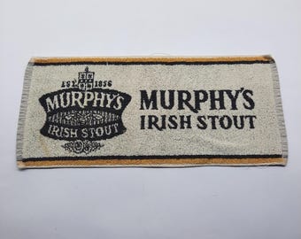 May include: A rectangular bar towel in off-white with black and gold trim. The towel features the black text Murphy's Irish Stout and a logo with the text Est. 1856, Murphys Irish Stout, and a crest design. The fabric has a textured, looped surface.