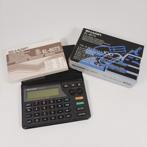 May include: A black Sharp EL-6073 Tel & Schedule 100 electronic organizer with a calculator keypad and buttons for functions like 'Fax', 'Remark', 'Secret', and 'Enter'. The organizer has a small LCD screen and is sitting on a white surface. The box for the organizer is in the background and is blue and white with the text 'Sharp EL-6073 Tel & Schedule 100' printed on it.