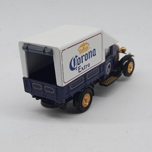 May include: A miniature vintage-style delivery truck featuring a dark blue cab and chassis with a white cargo box. The side of the white box displays the Corona Extra logo in blue and gold. The model includes gold-colored wheels and black tires.