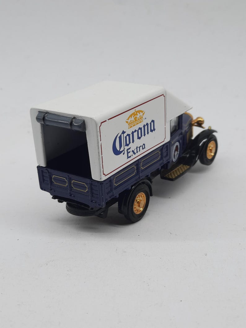 May include: A miniature vintage-style delivery truck featuring a dark blue cab and chassis with a white cargo box. The side of the white box displays the Corona Extra logo in blue and gold. The model includes gold-colored wheels and black tires.